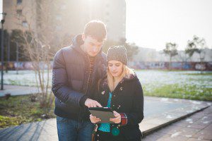 Knee figure of a couple of young beautiful caucasian woman and man strolling outdoor in the city back light, holding a tablet, looking downward and tapping the screen - love, technology, communication concept